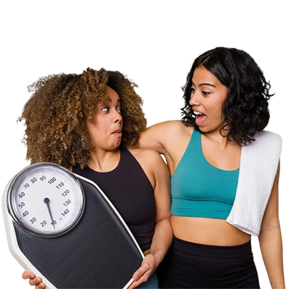 Two women in workout attire share a surprised, playful expression. One holds a large scale, the other has a towel draped over her shoulder. Black background.
