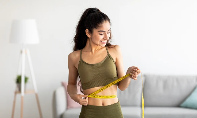 A smiling woman in a green workout outfit measures her waist with a yellow tape, standing in a bright living room. The scene conveys health and positivity.