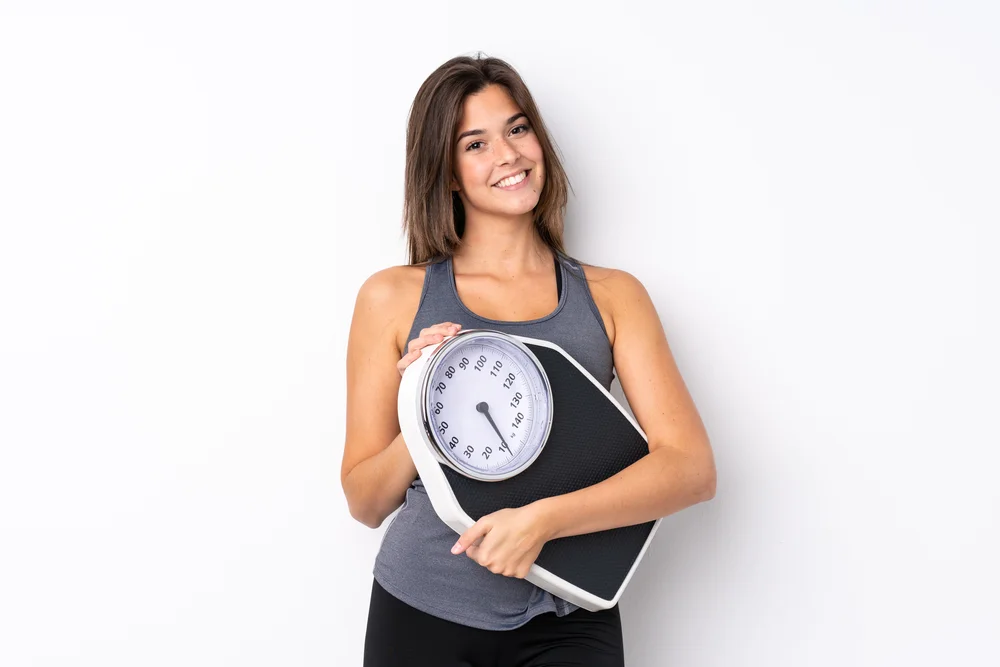 A woman in a gray tank top smiling while holding a bathroom scale.