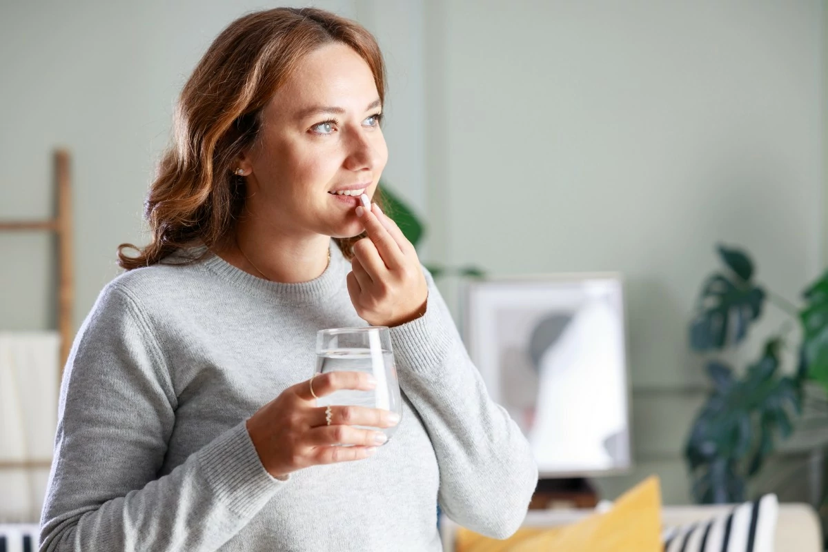 Woman in a gray sweater holds a glass of water and touches her lips thoughtfully. She stands in a bright room, creating a calm and pensive mood.
