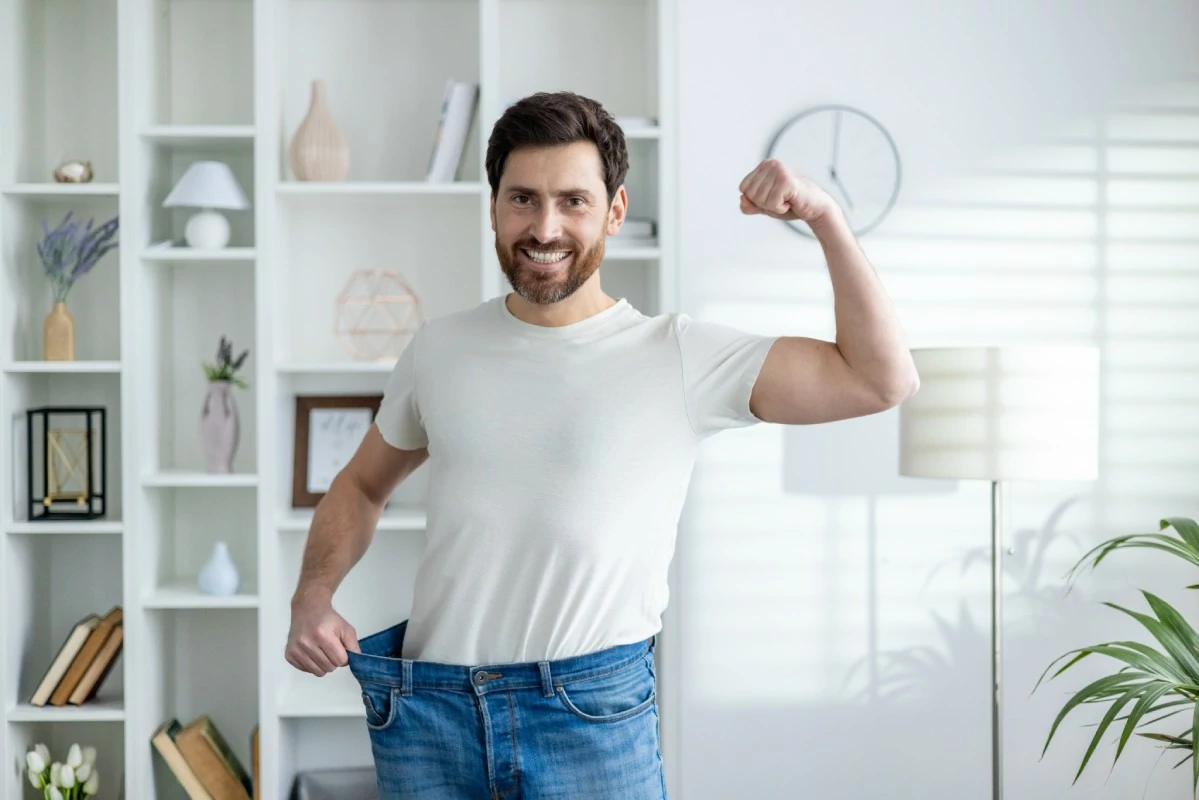 A smiling man in a white t-shirt shows off weight loss by holding out the waistband of oversized jeans. A bookshelf and lamp are in the bright room.